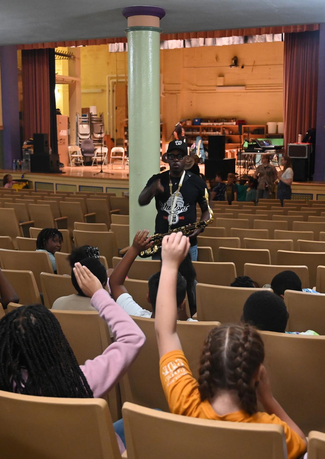 Students raise their hands as a saxophonist leads a session in a school auditorium