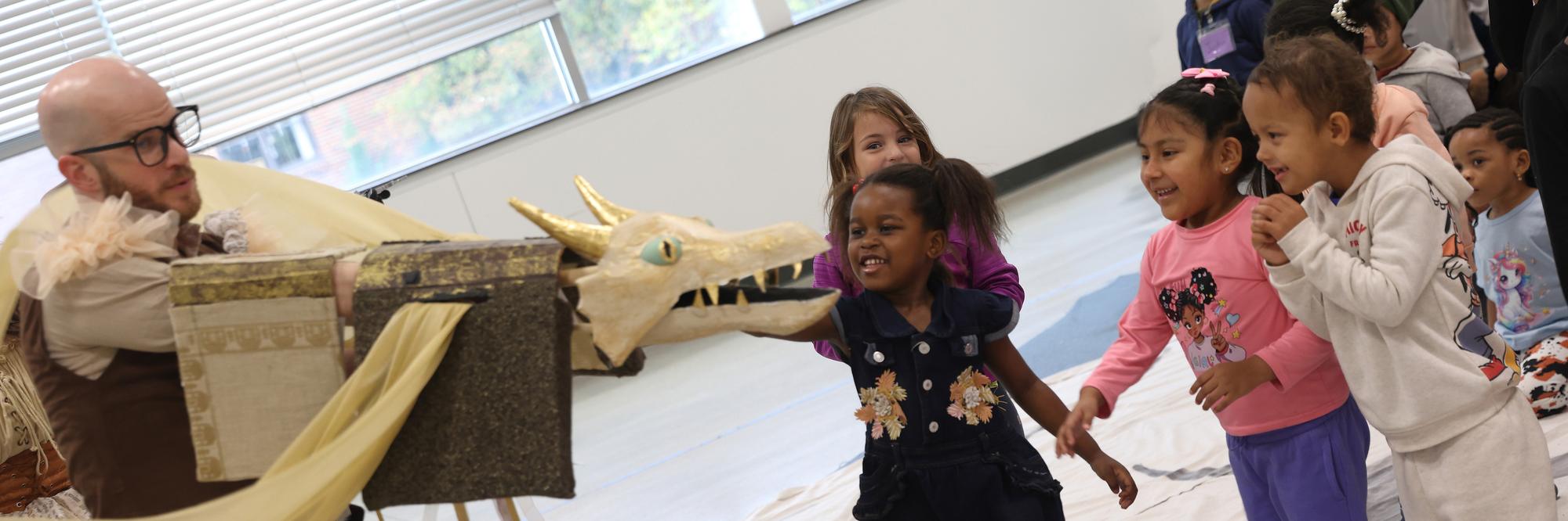 Preschoolers laugh as a teaching artist’s dragon puppet reaches toward them